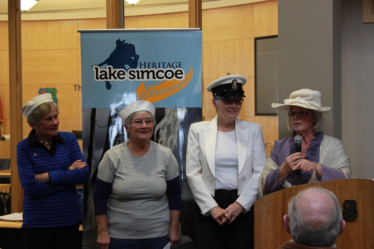 four women dressed in sailor hats and one in a bonnet stand behind a podium.