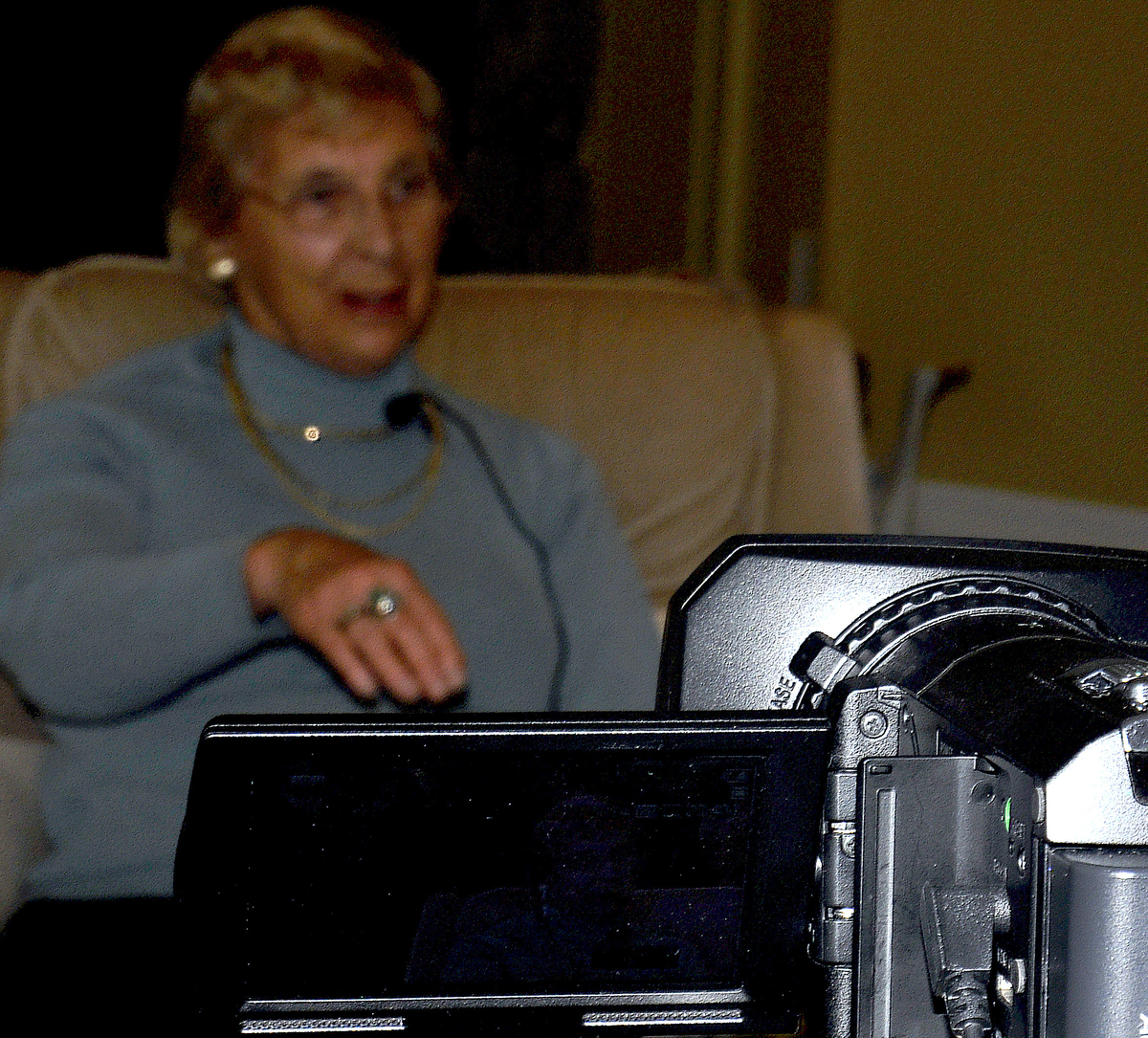 Video camera in foreground films woman in with short blonde hair, gasses in blue sweater