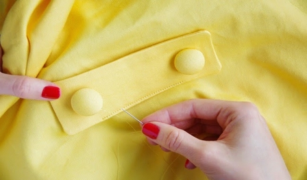 Image of a woman's hand with red fingernails sewing a button onto a yellow textile